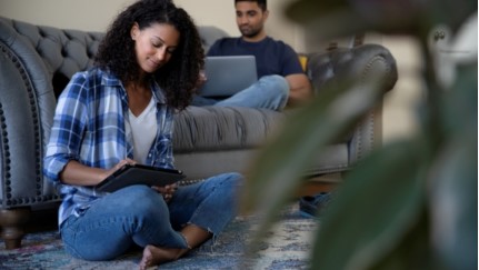 man sitting on a couch with a laptop and woman sitting on the floor with a tablet