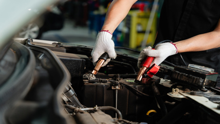 A person wearing gloves attaches black and red jumper cables to a car battery.