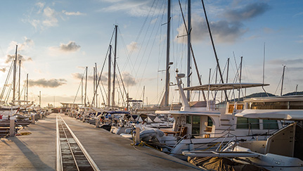 Sunset over a marina with sailboats and yachts docked along a long concrete pier.