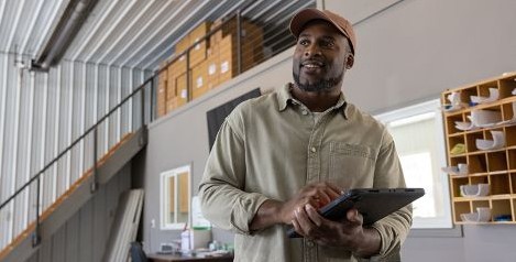 Man standing in a warehouse-style office holding a tablet with shelves and mail slots in the background.