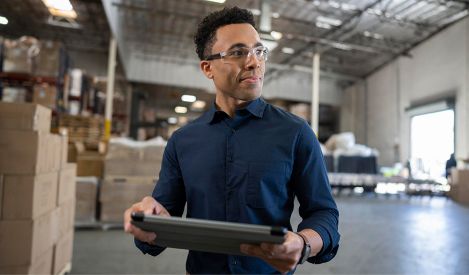 Man standing in a warehouse-style office holding a tablet with shelves and mail slots in the background.