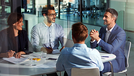 Four business colleagues are collaborating around a small oval table in a modern glass-walled office.