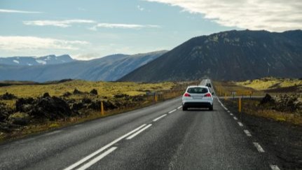 Car driving on a paved highway through a mountainous landscape under a partly cloudy sky.