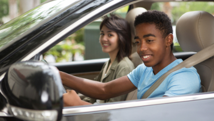A teenage boy sits in the driver’s seat of a car and checks his side mirror.