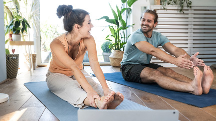 A young married couple laughs while practicing yoga at home in front of their laptop.