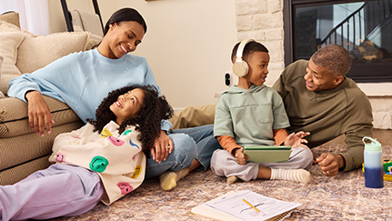 Parents smile while sitting on the living room floor with their two children, a grade-school boy wearing headphones and a tween girl resting her head on her mother’s lap.