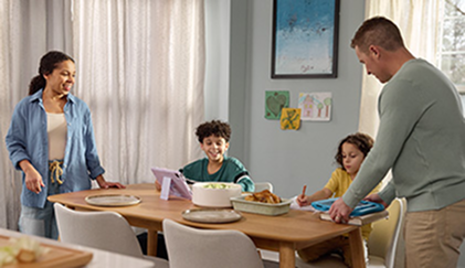 A family gathers around a table with a woman standing, a man serving food, and two kids seated with a tablet.