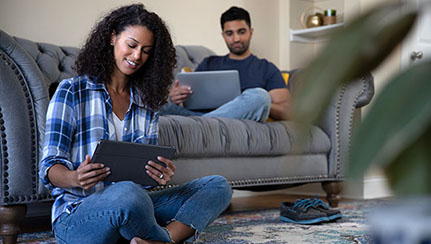man sitting on couch looking at his tablet and woman sitting on the floor looking at her tablet