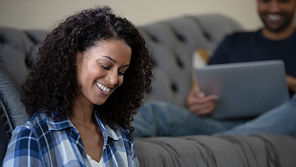 woman smiling while man sits in background on couch with laptop