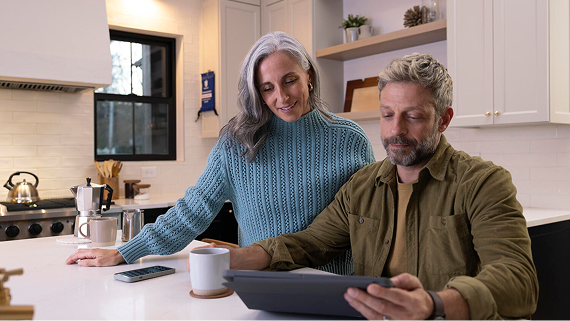 A smiling couple sits together in a bright kitchen, reviewing a tablet over coffee.