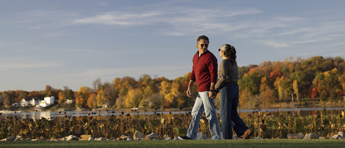 A retired couple takes an evening walk with a pond and fall foliage in the background.
