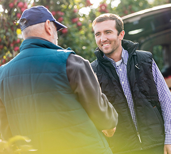 A farmer shakes hands with a buyer from a competing company in an orchard, as the buyer stands beside a van loaded with crates and holds a clipboard