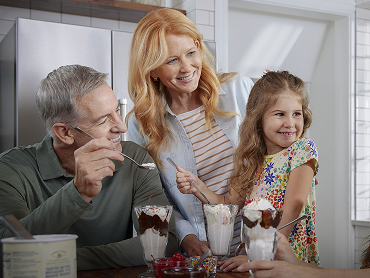 A retired couple smiles while sitting at their kitchen table eating ice cream sundaes with their young granddaughter.