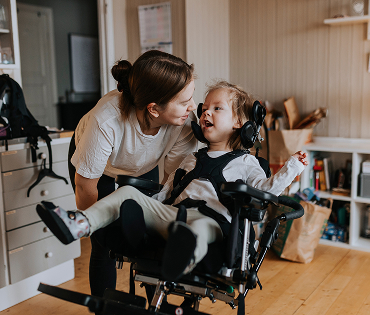 A female caregiver provides support to a smiling child in an activity chair in a home setting.