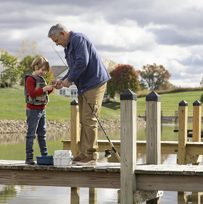 A retired man helps his grandson bait a hook while standing on a pier surrounding a small lake.