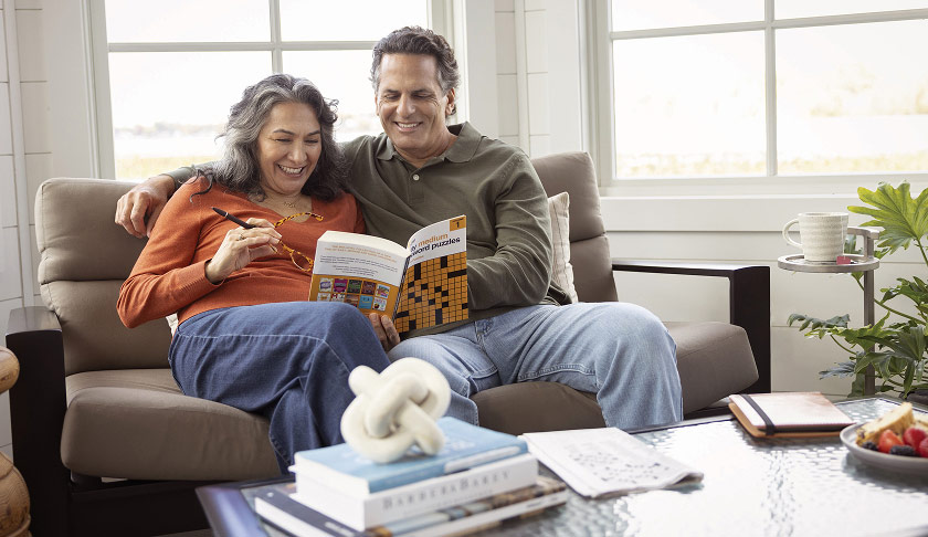 A retired couple sits together on their couch while laughing and completing a crossword puzzle.