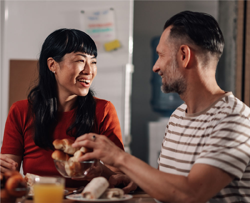 Man and woman sitting at a table, eating food and smiling at each other.