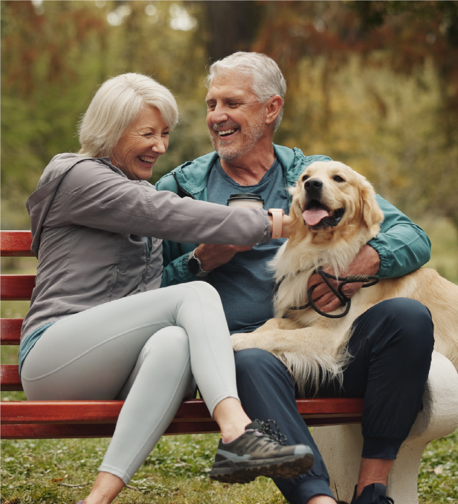 Couple in their 60s-70s sitting on a parch bench with their golden retriever and smiling.