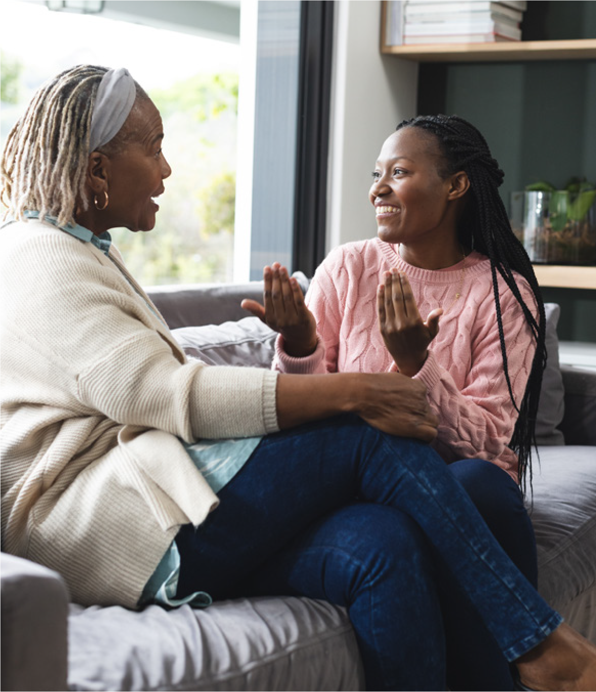 Mother and adult daughter sitting on a couch facing each other and smiling.
