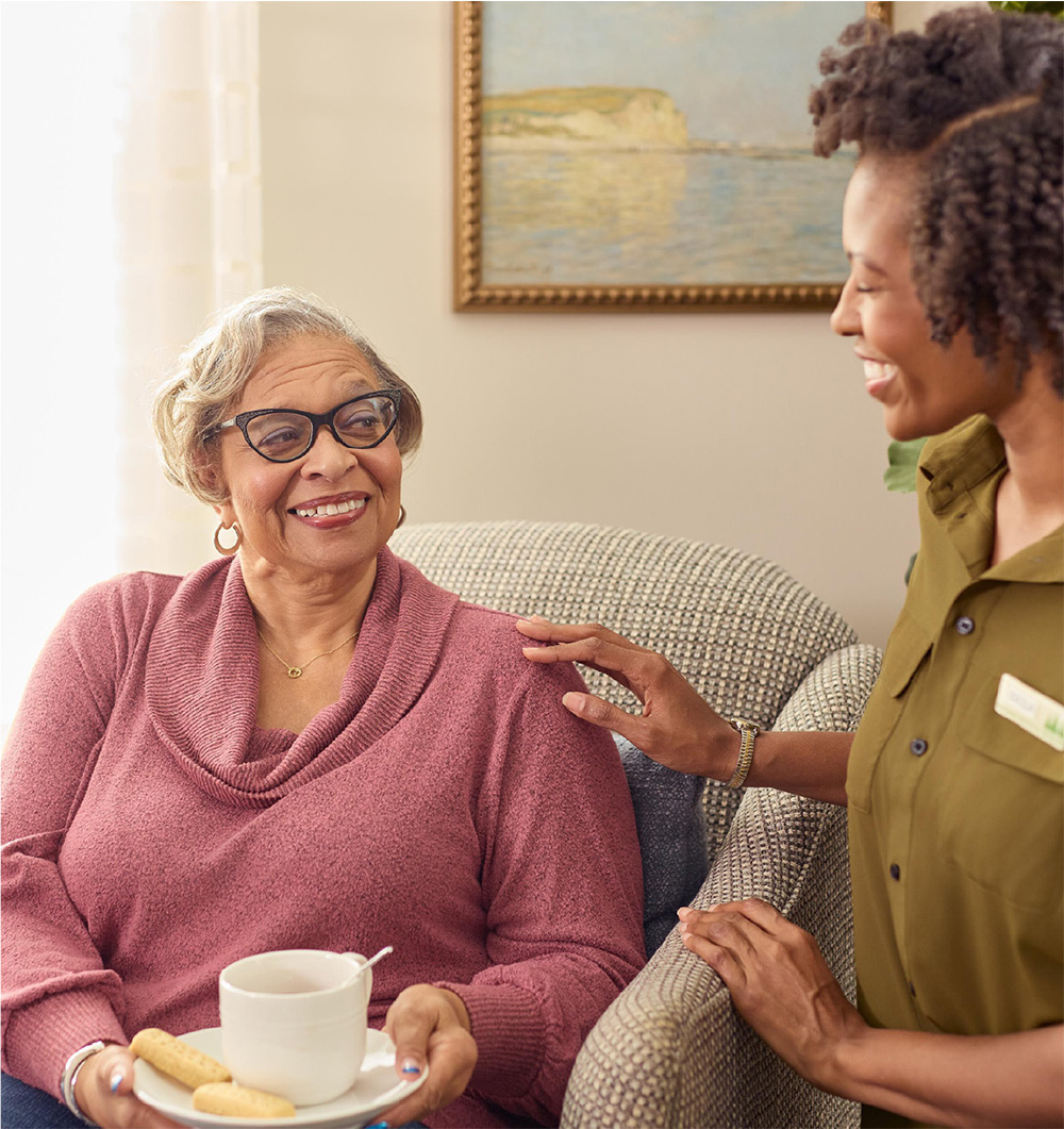 Woman in a nursing home sitting down and smiling at worker while drinking a cup of tea.