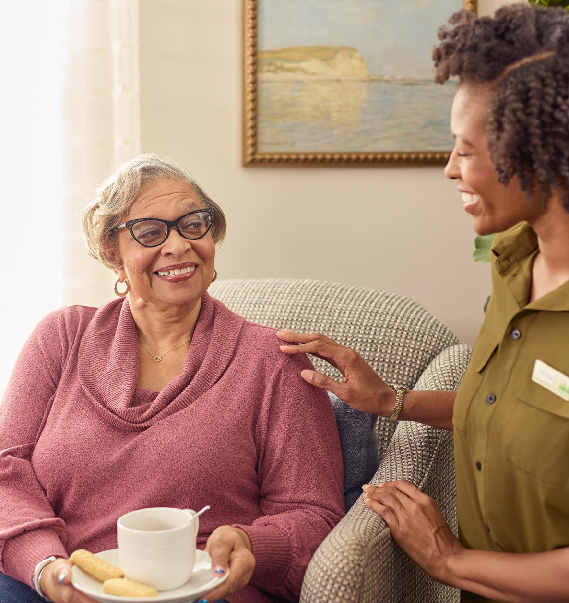 Woman in a nursing home sitting down and smiling at worker while drinking a cup of tea.