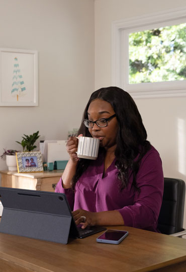 Woman in her mid 30s sitting at a desk on her tablet, drinking from a mug.