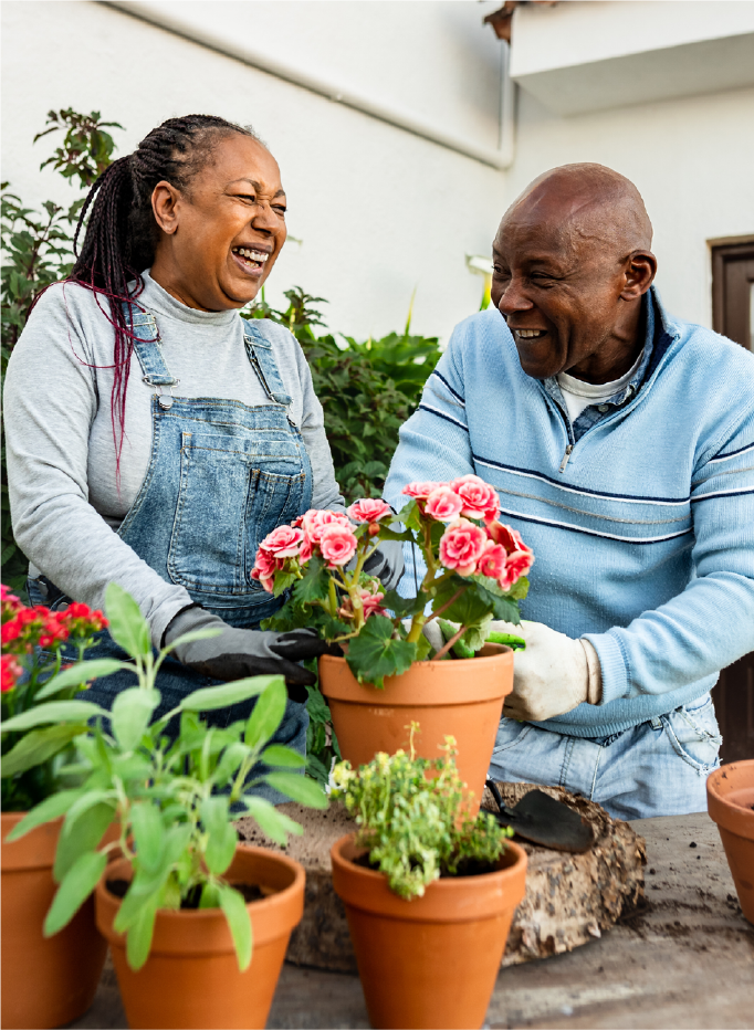 Couple in their 60s outside gardening and smiling while holding a pot of flowers.