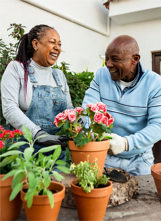 Couple in their 60s outside gardening and smiling while holding a pot of flowers.