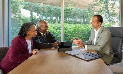 A senior couple and a financial planner seated at a wooden table in a modern office, engaged in a discussion with documents and a tablet on the table