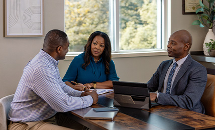 A senior couple and a financial planner seated at a wooden table in a modern office, engaged in a discussion with documents and a tablet on the table