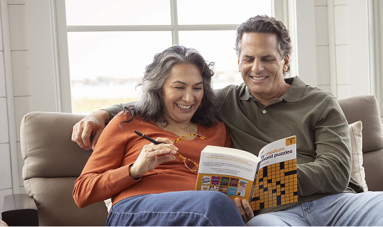 A retired couple sits together on their couch while laughing and completing a crossword puzzle.