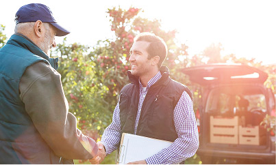 A farmer shakes hands with a buyer from a competing company in an orchard, as the buyer stands beside a van loaded with crates and holds a clipboard
