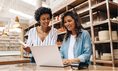 Two co-owners working together in a ceramic shop—one holding a clipboard, the other using a laptop at the counter, surrounded by shelves of pottery