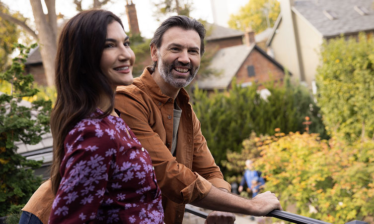 A middle aged couple smile while standing on their balcony.