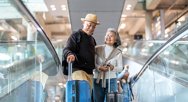 A retired couple rides an escalator at an airport with their young grandchildren. The grins on their faces show excitement about having time with the kids.