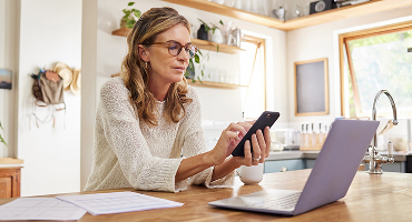 A middle-aged woman sits at her kitchen table referring to her phone, a laptop, and a pile of papers as she considers what to do with some inherited money.