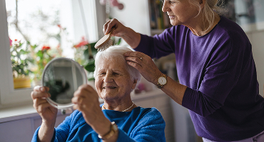 A woman in her 50s gently combs her mother's hair in her growing role as her mother's caretaker.
