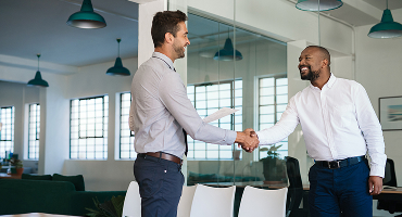 Two men in an office setting shake hands as one accepts the news of his promotion from the other
