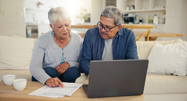 A couple approaching retirement age discusses their finances as they sit side by side on their sofa, leaning over a coffee table that holds their laptop computer and some papers.