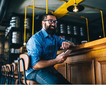 A business owner sits at a wooden counter in a modern café, holding a smartphone, with metal kegs and industrial piping in the background