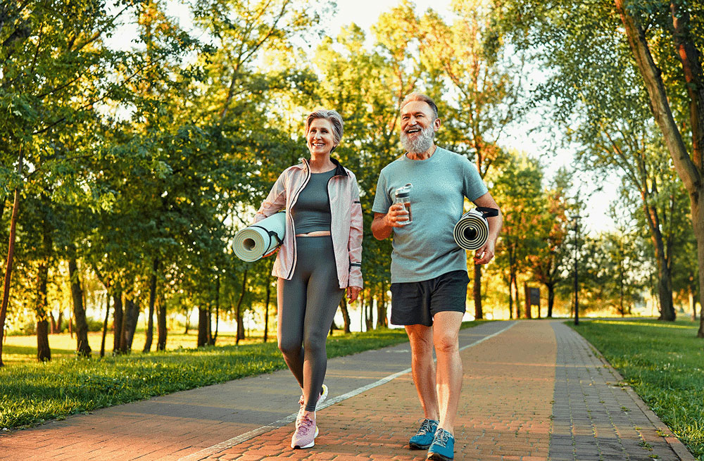 Two retirees walking on a tree-lined park path, carrying yoga mats and embracing a fulfilling retirement made possible by the security of protected lifetime income.