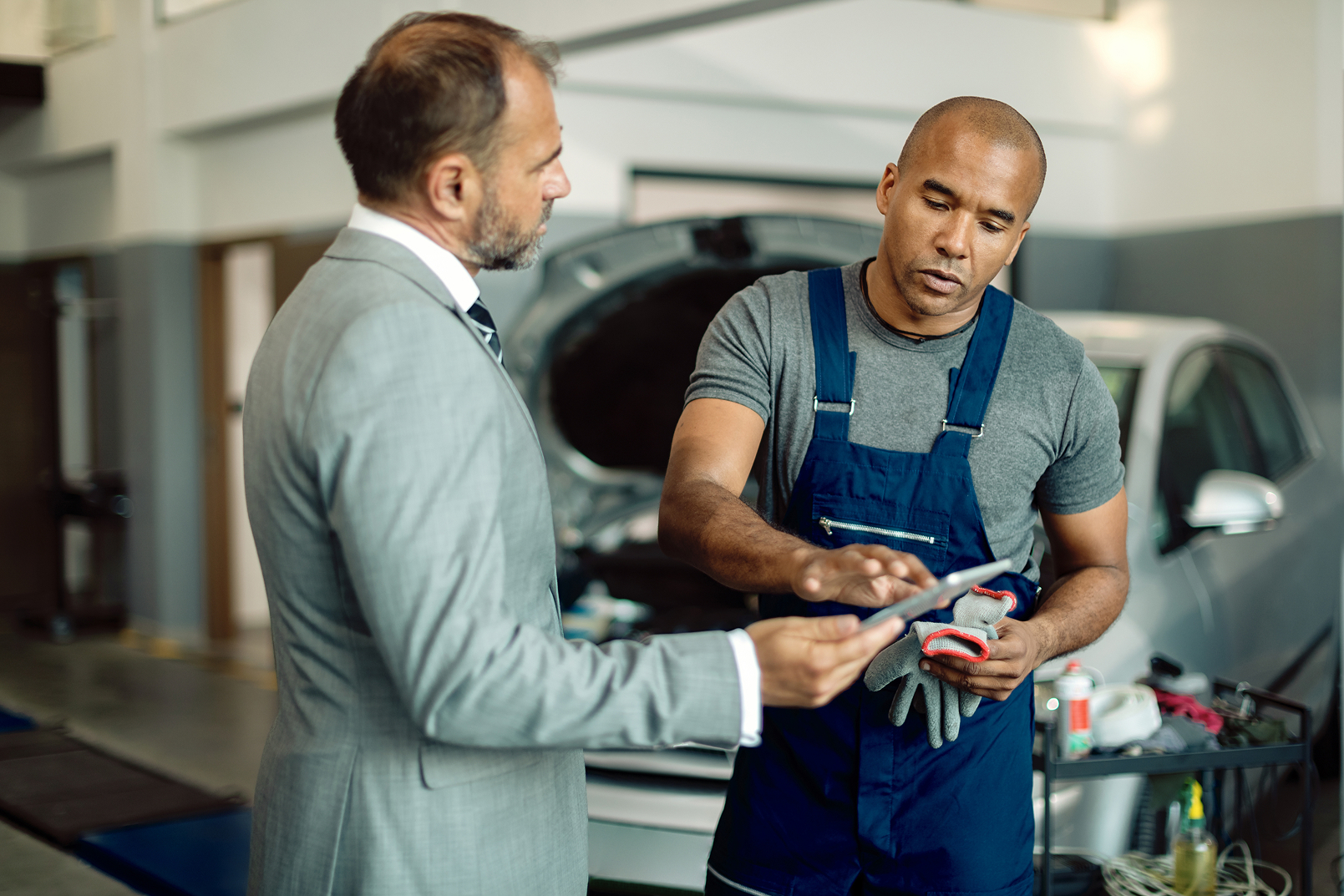 An auto shop owner discusses paperwork with their financial professional beside an open car hood.