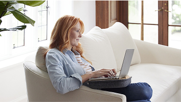 Middle-aged woman in a light blue denim shirt sits on a sofa in her living room researching her retirement income needs on her laptop.