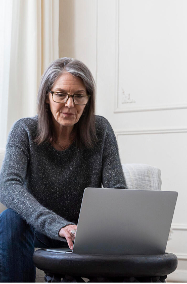 Person sitting on a sofa using a laptop on a small ottoman, with books and cushions nearby.