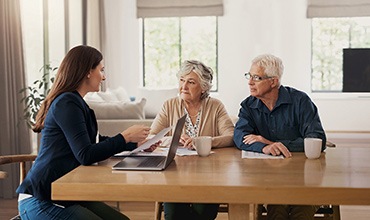 A senior couple and a financial planner seated at a wooden table at their home, engaged in a discussion with documents and a tablet on the table.