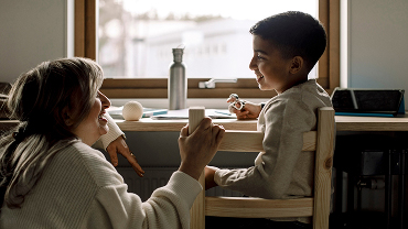 A mom kneels beside her child seated at a desk in front of a window.