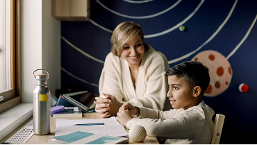 A mother and child siting at a desk covered with papers, a water bottle and a fidget.
