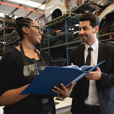 A business owner in a black apron discusses documents with a financial advisor in a suit inside an auto repair shop, surrounded by shelves of car parts and equipment