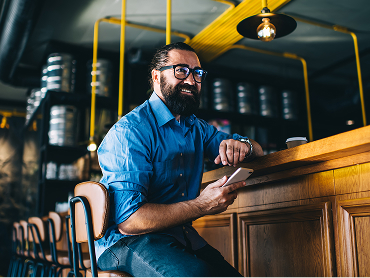 A business owner sits at a wooden counter in a modern café, holding a smartphone, with metal kegs and industrial piping in the background