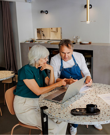A financial advisor and a shop owner discuss business over a laptop in a café.